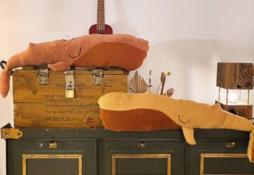 Zwei selbstgenähte Stillkissen in Form von Walen liegen auf großen Holzkisten, im Hintergrund eine Gitarre