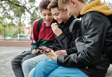 Drei Jungs sitzen mit Pausenbroten auf dem Schulhof und gucken auf ein Smartphone