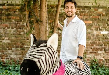 Mann im weißen Hemd mit Plüschzebra vor Baum und Mauer