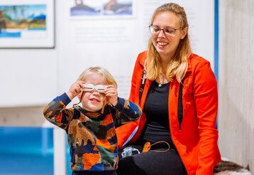 Kind hält sich eine Schneebrille vor die Augen, Frau sitzt dahinter