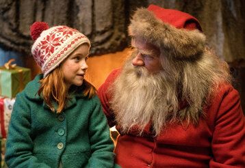 ein Mädchen mit Norweger-Mütze und grünem Mantel sitzt neben dem Weihnachtsmann und spricht mit ihm