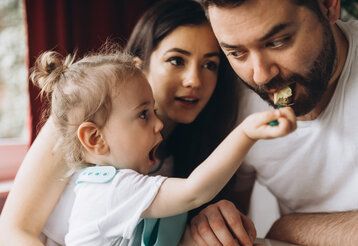Kind füttert Vater mit einem Löffel, die Mutter guckt zu
