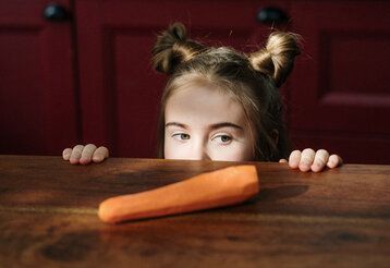 Mädchen guckt über die Tischkante, auf der eine Möhre liegt
