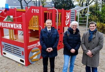 Drei Personen stehen auf einem Spielplatz, vor einem Spielgerät, das wie ein Feuerwehrauto aussieht