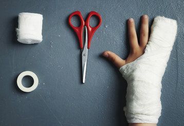 Mullbindenrolle, Kleberolle, Schere und verbundener Kinderarm von oben , dunkelgrauer Hintergrund