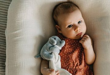 Baby liegt auf einem Kissen, ein Plüschelefant im Arm, guckt etwas kritisch in die Kamera
