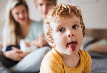 Kleiner Junge guckt in die Kamera und streckt die Zunge heraus, im Hintergund unscharf die Eltern mit einem Baby