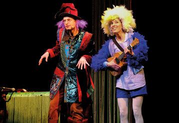 ein verkleideter Schauspieler und eine verkleidete Schauspielerin stehen auf einer Bühne, die Frau spiel Ukulele