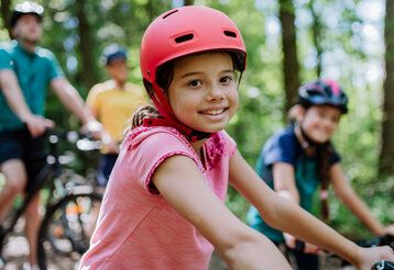 Mädchen mit rotem Fahrradhelm lächelt in die Kamera, im Hintergrund andere Radfahrer und Bäume, unscharf