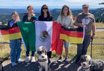 Eine Familie miteiner Austauschschülerin steht mit deutschen und mexikanischen Flaggen und zwei Hunden auf einem Berg