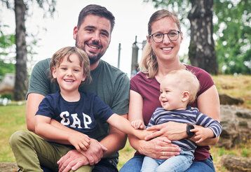 Vater und Mutter sitzen im Park, beide haben jeweils einen Sohn auf dem Schoß.