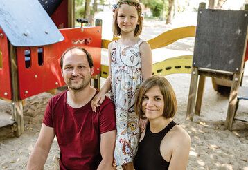 Vater und Mutter hocken im Sandkasten, Tochter steht mittig hinter ihnen