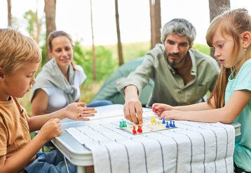 eine Familie spielt Mensch-ärgere-dich-nicht auf einem Campingtisch im Wald