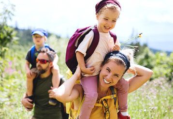 Familie wandert über eine Blumenwiese, Kinder bei Vater und Mutter huckepack