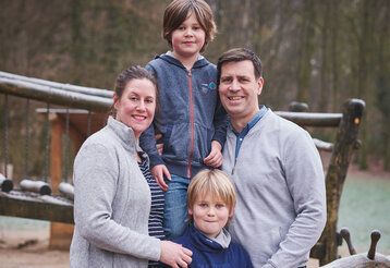 Mutter, Vater und zwei Jungs stehen zusammen auf einem Kinderspielplatz, im Hintergrund windertliche Bäume