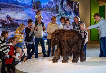 Besuchergruppe steht bei einer Figur eines Baby-Mammuts im Neanderthal Museum, der Museumsführer erklärt etwas