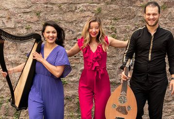 Zwei Musikerinnen und ein Musiker stehen mit Instrumenten vor einer Mauer