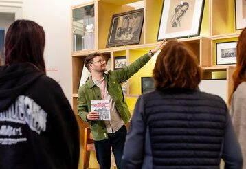 junger Mann erklärt etwas im Theatermuseum, er zeit auf ein Bild, Besucherinnen von hinten im Vordergrund