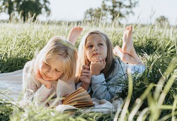 Zwei Mädchen liegen auf einer Decke im hohen Gras, eins liest in einem Buch