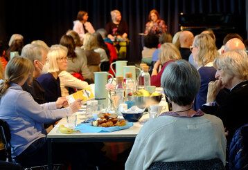 Leute sitzen zusammen an einem Tisch, auf dem Frühstück bereitsteht, im Hintergrund eine Bühne, auf der sich drei Frauen unterhalten