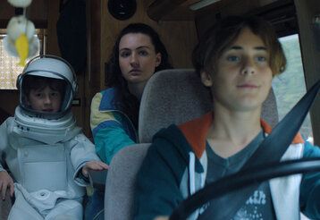 ein als Astronaut verkleidetes Kind sitzt mit einem großen Mädchen in einem Wohnmobil, im Vordergrund ein großer Junge, der das Fahrzeug fährt