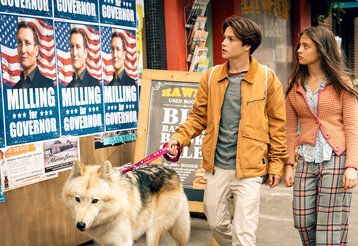 Szene aus dem Film Woodwalkers 2, ein Junge undein Mädchen laufen mit einem Hund an Wahlplakaten vorbei