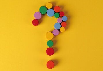 ein Fragezeichen, das aus bunten, runden Holzklötzchen gelegt ist, auf gelbem Hintergrund