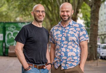 Porträt von Frank Walber und Markus Feger, im Hintergrund ein Platz in Düsseldorf mit Bäumen