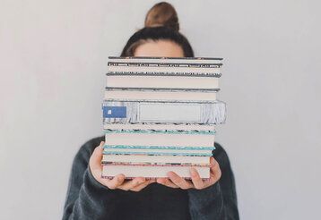 Frau hält einen Bücherstapel in die Kamera, der ihr Gesicht verdeckt