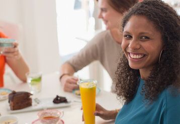 Frau sitzt an einem Esstisch mit einem Glas Orangensaft, im Hintergrund zwei Frauen, die sich unterhalten