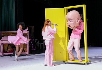 drei Schauspieler:innen in rosa Kleidung auf der Bühne, eine gelbe Tür auf der Bühne, ein Schauspieler hält sich eine große Baby-Maske vors Gesicht.