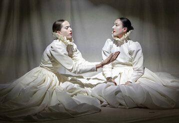 Zwei Frauen in weißen historisch anmutenden Kleidern sitzen auf dem Boden vor weißem Vorhang