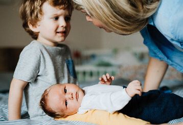 Baby liegt auf einer Decke und guckt in die Kamera, Bruder sitzt dabei und wirkt unzufrieden, Mutter spricht mit ihm