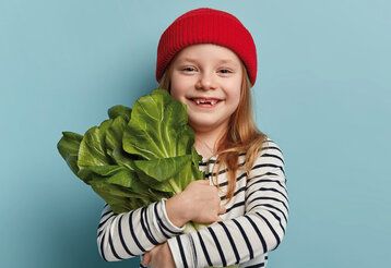 Mädchen mit Ringel-Shirt und roter Mütze hält Salat im Arm, vor blauem Hintergrund
