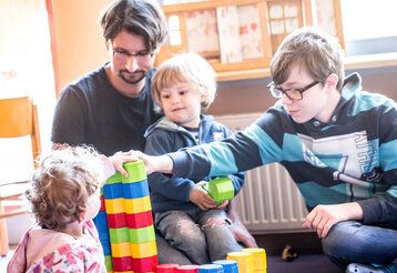 Ein Erzieher und ein Junge sitzen in einer Kita und spielen mit zwei Kleinkindern
