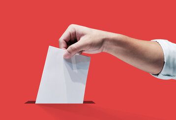 eine Hand steckt einen Wahlzettel in einem Schlitz, der Hintergrund ist rot