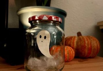 Einmachglas mit einem kleinen Papiergespenst, im Hintergrund Kürbisse und ein Übertopf mit einer Pflanze
