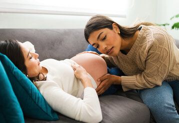 Schwangere Frau liegt auf einem Sofa und wird von einer Hebamme untersucht.