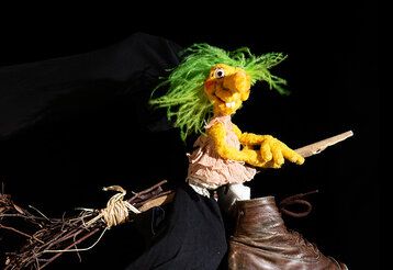 Die Puppe Hexe Hilda reitet vor schwarzem Hintergrund auf ihrem Besen.