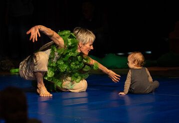 eine mit Efeu behangene Tänzerin kniet neben einem Kleinkind