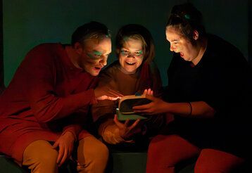 ein Schauspieler  und zwei Schauspielerinnen sitzen im Dunklen zusammen und gucken auf etwas leuchtendes