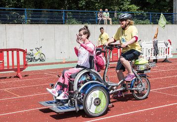 Special Petit Départ für Kinder mit Handicap im Rather Waldstadion, Frau fährt mit speziellem Rad, auf dem ein Rollstuhl installiert ist, auf dem ein Mädchen sitzt, auf einer Aschebahn