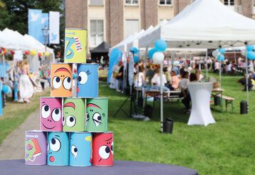 Jahresfest inder Kaiserswerther Diakonie, Biertische und Pavillons im Garten der Diakonie mit Besucher:innen, im Vordergrund gestapelte Blechdosen mit lustigen Gesichtern