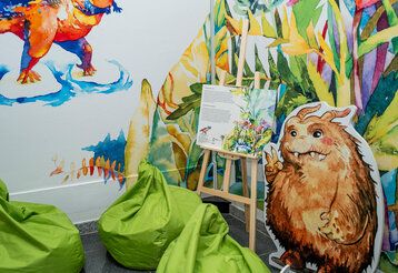 Ecke eines Ausstellungsraumes im Oberschlesischen Landesmuseum mit grünen Sitzsäcken, einem Aufsteller in Form eines kleinen Monsters und eine Staffelei, auf der ein aufgeschlagenes Buch liegt