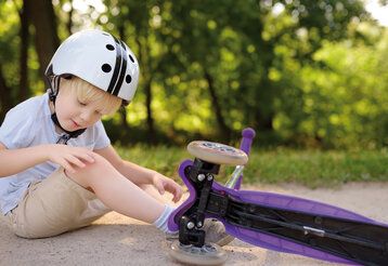 Kleiner Junge mit Helm sitzt auf einem Weg und betrachtet sein Knie, er ist mit Kickboard hingefallen. Das lila Kickboard liegt neben ihm. Im Hintergrund Bäume.