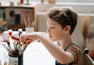 Junge sitzt am Tisch mit Malutensilien und nimmt einen Pinsel aus einem Becher mit Pinseln