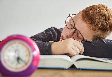 ein Junge schläft auf einem Buch, im Vordergrund steht ein Wecker