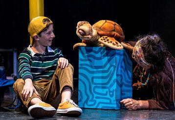Junge sitzt auf dem Boden und spricht mit Schildkrötenpuppe daneben sitzt die Puppenspielerin