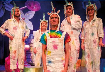 Sechs Schauspieler:innen mit weißen Latzhosen und Einhörnern auf dem Kopf auf Bühne
