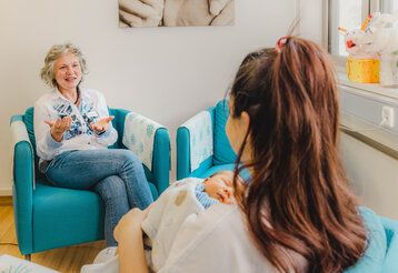 Zwei Frauen im Gespräch, eine ältere spricht, eine jüngere sitzt iht mit Neugeborenem gegenüber, dem Betrachter mit dem Rücken zugewandt.